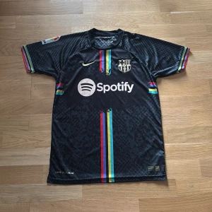 FC Barcelona Nike tröja - Snygg svart FC Barcelona matchtröja från Nike med Lewandowski och nummer 9 på ryggen. Tröjan har färgglada ränder i rött, blått och gult, korta ärmar och Dri-FIT-material som andas. Spotify-logga på bröstet och UNHCR-tryck på ryggen.