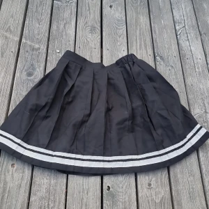 Svart plisserad kortkjol med vita ränder - Snygg svart plisserad kortkjol med två vita ränder längst ner. Kjolen har en klassisk tennisstil och resår i midjan. Saknar storlekslapp men skulle säga XXS-XS. 