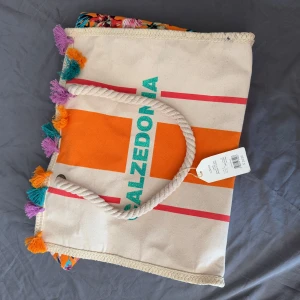 Calzedonia strandväska med tofsar - Stor strandväska från Calzedonia i beige bomull med orange och röda ränder, grönt tryck och färgglada tofsar i lila, turkos och orange. Rejäla rep-handtag och tropiskt mönstrat foder på sidorna. Perfekt för stranden och rymmer allt du behöver.