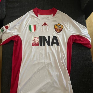 AS Roma Totti 10 bortatröja Kappa - Snygg AS Roma bortatröja från Kappa med Totti 10 på ryggen. Tröjan är vit med röda detaljer på sidor och ärmar, klubbmärke och sponsortryck framtill. Klassisk passform och tillverkad i lätt, glansigt material som andas. Perfekt för dig som älskar italiensk fotboll och legenden Totti.