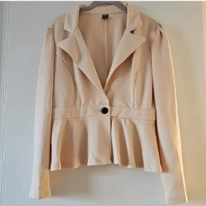 Beige kavaj med volang från SHEIN - Snygg beige kavaj från SHEIN i storlek M. Jackan har markerad midja, volang nedtill och en stor krage. Stängs med en knapp framtill och har långa ärmar. Perfekt för dig som vill ha en chic och feminin look.