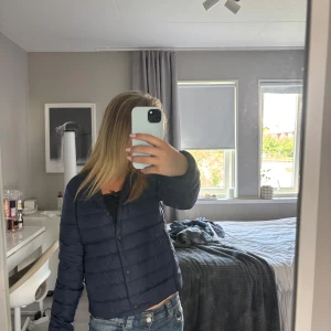 Mörkblå dunjacka från Uniqlo  - Säljer en stilren mörkblå dunjacka från Uniqlo. Väldigt bra på hösten eller när det börjar bli kallare Jackan är lätt, har rund halsringning och knäpps med knappar framtill. Den är quiltad och har en clean look utan krage, perfekt för lager-på-lager. Kostar egentligen 600kr, (inte använd)