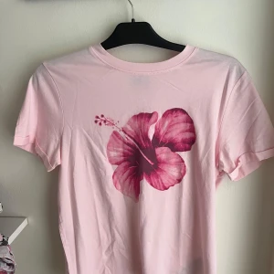 Ljusrosa t-shirt med blomtryck FB Sister - Ljusrosa t-shirt från FB Sister med en stor rosa hibiskusblomma tryckt framtill. Klassisk rund halsringning och korta ärmar. Skön regular fit i mjuk bomull, perfekt för en chill och färgglad look.