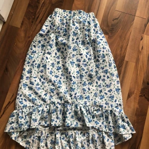 Vit blommig långkjol med volang - Supersöt långkjol i vitt med blått blommönster och gröna detaljer. Kjolen har elastisk midja och bred volang nertill som ger en romantisk och somrig vibe. Perfekt för dig som gillar en feminin och avslappnad stil.