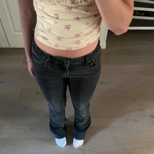 Svarta lågmidjade bootcut jeans - Säljer ett par svarta lågmidjade jeans med bootcut passform. Jeansen har snygga detaljer med dragkedjor och fickor framtill. Perfekta för dig som gillar en klassisk men edgy stil. Materialet är jeans och färgen är mörkgrå till svart. Jag på bilden är 160 cm