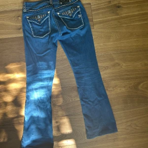 Blåa Miss Me bootcut jeans med detaljer - Snygga blå bootcut jeans från Miss Me med kontrastsömmar och blingade bakfickor med nitar och stenar. Jeansen har coola detaljer som broderier och metallknappar samt ett läderpatch bak i midjan. Perfekt för dig som gillar statement-jeans!