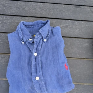  Ralph Lauren skjorta  - Snygg blå skjorta i linne från Ralph Lauren med korta ärmar och klassisk krage. Skjortan har vita knappar och den ikoniska röda broderade loggan på bröstet. Perfekt för en chill och stilren look.