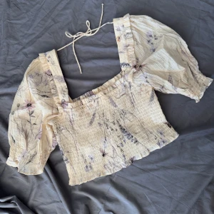 Blommig croppad blus med puffärm - Superfin ljusbeige croppad blus med blommigt mönster i lila från H&M. Blusen har puffiga ärmar, smockad kropp och volangdetaljer vid axelbanden. Knytband i nacken för extra söt look. Perfekt för vår och sommar!