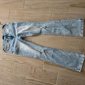 True Religion ljusa bootcut jeans - Säljer ett par ljusblå bootcut jeans från True Religion med slitningar och trasiga detaljer. Jeansen har klassiska bakfickor med röd kontrastsöm och logopatch. Materialet är jeans i bomull och passformen är loose med låg midja. Från Nelly