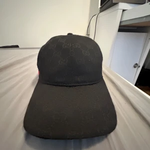Svart keps från Gucci med GG-mönster - Snygg svart keps från Gucci med diskret GG-monogram över hela kepsen. Klassisk böjd skärm och justerbar rem bak i skinn. Sidan har den ikoniska gröna och röda randen. Perfekt accessoar för dig som gillar lyxiga detaljer.