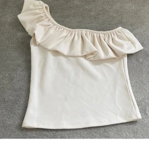 Beige oneshoulder volangtopp - Supersöt beige topp med volang längs en axel och oneshoulder-design. Perfekt för dig som vill sticka ut med en feminin och trendig look. Toppens material känns mjukt och följsamt mot huden. 