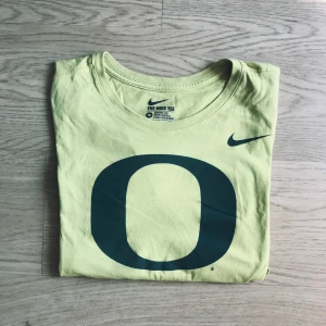 Gul Nike t-shirt med stor O-logga - Snygg gul t-shirt från Nike med stor mörkgrön O-logga på bröstet och klassisk Nike Swoosh. Modellen är kortärmad och i 100% bomull, perfekt för dig som gillar sportig och clean stil. Athletic cut för en schysst passform.