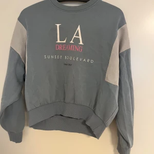 Blå sweatshirt LA Dreaming Divided XS - Blå sweatshirt från H&M Divided i storlek XS med trycket 'LA Dreaming Sunset Boulevard' framtill. Tröjan har ljusgrå paneler på ärmarna, ribbade muddar och avslappnad passform. Perfekt för en chill och trendig look.