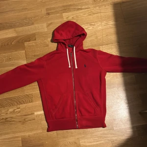 Röd hoodie från Polo Ralph Lauren - Röd hoodie från Polo Ralph Lauren med dragkedja, vita snören i huvan och klassisk blå logga på bröstet. Tröjan har känguruficka framtill och är tillverkad i mjuk bomull. Perfekt för dig som gillar en stilren och sportig look.