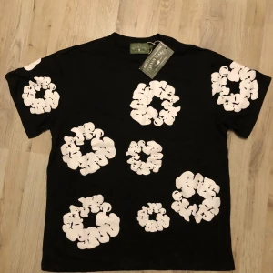 Svart Denim Tears T-shirt - En helt ny Svart t-shirt med tags från Denim Tears med stora vita blommiga tryck över hela plagget, både fram och bak. Klassisk passform med rund hals och korta ärmar. Skön bomullskvalitet och unik design som sticker ut.