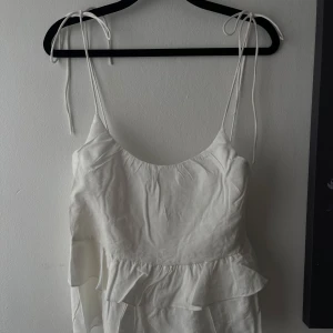 Vit volangtop med knytband - Supersöt vit linnetopp med tunna axelband som knyts upptill och volangdetaljer både fram och bak. Snyggt flowy fall och lätt att matcha med jeans eller kjol.