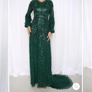 Balklänning/Festklänning - Magisk långklänning från Shimmidresses i djupgrönt med glittrande paljetter över hela tyget. Klänningen har långa ärmar, markerad midja och en snygg rosett baktill. Den har en elegant släp och är perfekt för bal eller fest. Materialet känns lyxigt och skimrande. Aldrig använd, prislapp kvar. 