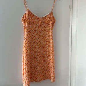 Blommig orange kort sommarklänning - Söt kort klänning med orange och vita blommor, smala axelband och liten knytning framtill. Perfekt för varma dagar och har en lätt, flowy passform. Materialet känns mjukt och luftigt.