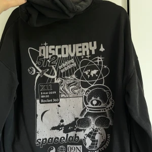 Svart NASA hoodie med tryck - Svart hoodie med stort grafiskt print på ryggen med rymdtema, astronaut-nalle och text. Hoodien har huva med snörning, känguruficka och långa ärmar. Lite streewear vibes