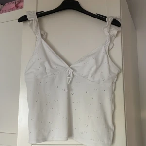Vit linnetopp med knyt från H&M Divided - Supersöt vit linnetopp från H&M Divided med broderade hålmönster och volangiga axelband. Toppen har v-ringning och knyt fram samt snygg öppen rygg med dubbla knytband. Perfekt för varma dagar.