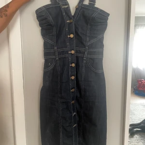Mörkblå jeansklänning med knappar - Snygg mörkblå jeansklänning med markerade sömmar och knappar hela vägen framtill. Axelbandslös design med breda avtagbara axelband och coola fickdetaljer. Perfekt för dig som gillar denim och vill ha en klänning med edge.