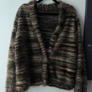 Randig fluffig kofta - Supermysig och fluffig kofta med breda ränder i jordnära färger som brun, beige och grön. Koftan är stickad, har lång ärm, v-ringning och knäpps med två knappar framtill. Perfekt för lager-på-lager och chill vibes.