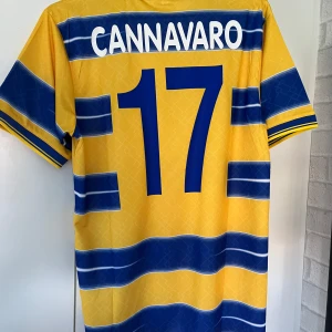 Parma Cannavaro 17 retro fotbollströja - Retro Parma AC fotbollströja med Cannavaro 17 på ryggen. Klassisk gul och blå design med breda horisontella ränder, klubbmärke och Parmalat-logga på bröstet. Tillverkad i lätt och ventilerande polyester, perfekt för dig som älskar vintage och italiensk fotboll. Helt ny