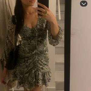Zara klänning  - Supersnygg kort klänning med grönt och svart mönster, v-ringning och rynkade detaljer framtill. Klänningen har långa ärmar och volangkant nertill för en extra feminin touch. Perfekt för dig som vill sticka ut med en trendig look.
