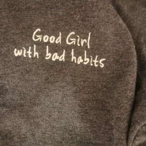 Mörkgrå croppad hoodie med text - Mörkgrå croppad hoodie med huva och snörning. På bröstet står det 'Good Girl with bad habits' i vit brodyr. Tröjan har långa ärmar och är tillverkad i mjukt bomullsmaterial. 
