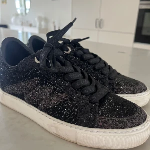Svarta glittriga sneakers Zadig & Voltaire - Svarta sneakers från Zadig & Voltaire med glittrig yta och dekorativ stjärna på sidan. Skorna har svarta snören med logga och vit platt sula. Perfekta för dig som vill sticka ut med lite extra bling i vardagen.