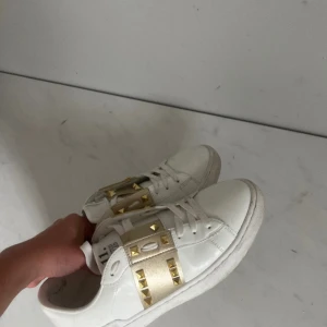 Valentino Rockstud vita sneakers - Snygga vita Valentino Rockstud sneakers med guldiga nitar och breda band i satin över vristen. Skorna har klassisk rund tå, platt sula och snörning. Perfekta för dig som vill sticka ut med exklusiva detaljer och lyxig känsla. Materialet är skinn med inslag av satin. 