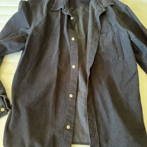 Mörkgrå manchester overshirt - Snygg mörkgrå overshirt i manchester med klassisk krage, bröstficka och vita knappar. Perfekt att slänga över en t-shirt för en avslappnad look. Långärmad och loose fit för skön känsla.