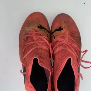 Rosa fotbollsskor från Puma - Säljer ett par snygga fotbollsskor från Puma i en cool rosa färg med svarta Puma-loggor på sidan och ovansidan. Skorna har snörning och en stickad ovandel för skön passform. Sulan har dobbar som ger bra grepp på planen. Perfekt för dig som vill sticka ut på matchen!