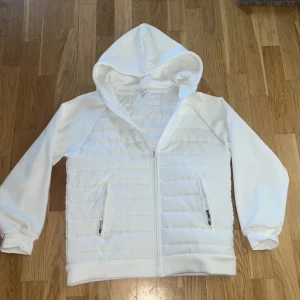 Vit pufferjacka med huva i storlek M men kan också passa S - Snygg vit pufferjacka med huva och dragkedja framtill. Jackan har quiltade partier på framsidan och två fickor med dragkedja. Perfekt för dig som gillar en clean och sportig stil. Storlek M men passar också storlek S och cardiganen är aldrig använd.
