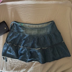 Volangkjol i jeans, strl S - Säljer en kort jeanskjol med volanger i mörkblå denim. Kjolen är tillverkad i 100% bomull och har en avslappnad passform med snygga sömmar och rynkade detaljer. Perfekt för dig som gillar en chill och trendig look.