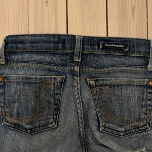 Rock & Republic bootcut jeans blå - Snygga blå jeans från Rock & Republic med bootcut passform. Klassisk femficksmodell och broderade bakfickor. Jeansen har låg midja och är tillverkade i jeansmaterial med en cool tvättad look. Jeansen är i storlek 24. Tyvärr har jeansen ett rev nedanför rumpan, och materialet på jeansen har blivit ganska tunt 🫶