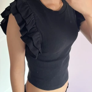 Svart volangärmad topp från Zara - Supersnygg svart topp från Zara med ribbad struktur och volangdetaljer på axlarna. Modellen är croppad och ärmlös, vilket ger en trendig och cool look. Perfekt att styla med jeans eller kjol för en extra touch.