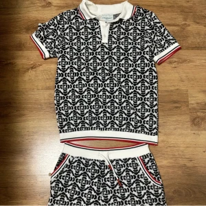 Casablanca Set. - Casablanca t-shirt med vit krage och knappslå, svartvit grafiskt mönster över hela plagget. Ribbade muddar med röda och svarta ränder på ärmar och nederkant. Snygg och sportig stil, perfekt för tennis eller chill. Materialet känns som bomull. Shorts tillkommer.