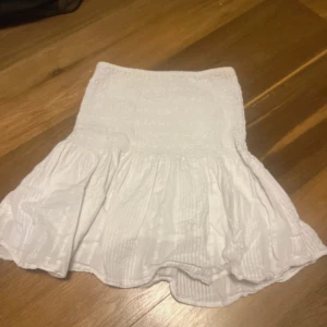 Vit kortkjol med volang kan också användas som topp  - Supersöt vit kortkjol med volangkant och smockad midja. Kjolen har ett diskret randigt mönster i tyget och är gjord i lätt bomull som känns luftig och fräsch. Perfekt för varma dagar och ger en chill vibe till din outfit.