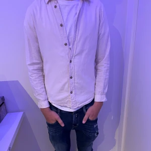 Vit långärmad linneskjorta skjorta  - Beige linneskjorta med klassisk krage och bruna knappar. Modellen på bild är 1,75cm lång och väger 56 och den sitter slim på modellen. Märket är Jack and Jones. 