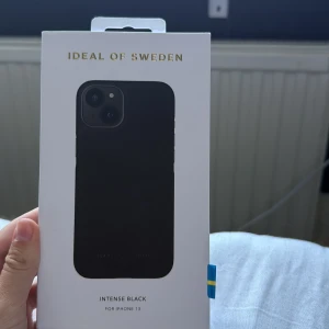 Svart mobilskal Ideal of Sweden - Snyggt svart mobilskal från Ideal of Sweden, designat för iPhone 13. Skalets stil är minimalistisk och elegant med en matt finish och diskret logga. Perfekt för dig som vill ha en clean look på mobilen. Helt oanvänt
