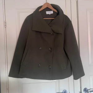 Olivgrön skepparkavaj från Bubbleroom - Snygg olivgrön skepparkavaj från Bubbleroom med bred krage och dubbelknäppning. Jackan har raka, långa ärmar och två fickor framtill. Perfekt för dig som vill ha en klassisk och stilren look under höst och vinter.