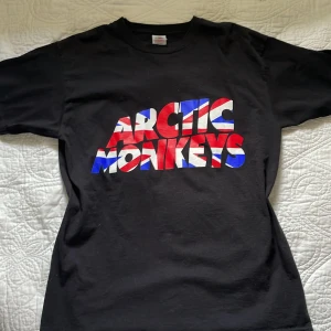 Arctic Monkeys t-shirt svart M - Svart t-shirt från Fruit of the Loom med Arctic Monkeys-tryck på både fram- och baksidan. Framsidan har bandets logga i rött, vitt och blått med brittisk flaggstil, medan baksidan har vit logga med soundwave-design. Klassisk rund hals och korta ärmar. Perfekt för fans!