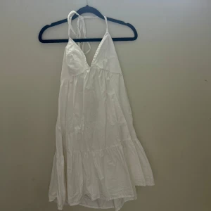 Vit omlottklänning från BikBok - Säljer en vit omlottklänning från BikBok i storlek M. Klänningen har smala axelband som knyts i nacken, v-ringad front och volangdetaljer nedtill. Den är ärmlös och har en luftig passform, perfekt för varma dagar. Materialet är mjuk bomull.