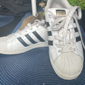 Adidas Superstar vita sneakers strl 38 - Klassiska Adidas Superstar sneakers i vitt skinn med svarta ränder och logga på hälen. Skorna har snörning, rund tå och den ikoniska gummitåskyddet. Perfekta för dig som gillar en tidlös streetstil.