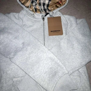 Grå zip-hoodie från Burberry M - Snygg ljusgrå hoodie från Burberry med dragkedja och klassiskt rutigt foder i huvan. Jackan har snörning i huvan, ribbade muddar och en clean look. Perfekt för dig som gillar exklusiva streetwear vibes och vill ha något som sticker ut.