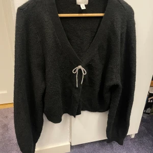 Svart stickad kofta med rosett från H&M - Mysig svart stickad kofta från H&M i storlek L (passar mig som vanligtvis har s/m, kortare än man tror). Koftan har v-ringning, långa ärmar och en glittrig rosett framtill. Stängs med knappar och har en loose passform som ger en chill vibe. Perfekt för att addera lite extra stil till din outfit.