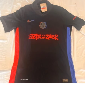 FC Barcelona x Cactus Jack Nike tröja - Unik svart FC Barcelona fotbollströja i samarbete med Cactus Jack och Nike. Tröjan har röda och blå sidopaneler, tryckt Cactus Jack-grafik på bröstet och UNHCR-logga på ryggen. Broderat klubbmärke och Nike-logga framtill. Tillverkad i lätt, tekniskt material.