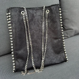 Svart axelväska med kedja och nitar - Snygg svart axelväska i mocka med silverfärgade kedjor som axelrem och coola nitar längs sidorna. Väskan har dragkedja upptill och ett rymligt fack. Perfekt för dig som gillar en edgy och stilren look.Fläck på baksidan, var god se sista bilden. Från Zara 