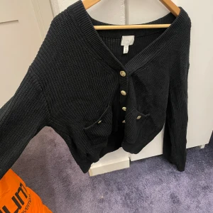 Svart stickad kofta från H&M med guldknappar - Svart stickad kofta från H&M i storlek S. Koftan har snygga guldknappar framtill och två fickor med dekorativa knappar. Modellen är v-ringad och har långa ärmar. Perfekt för dig som gillar klassisk stil med en twist.